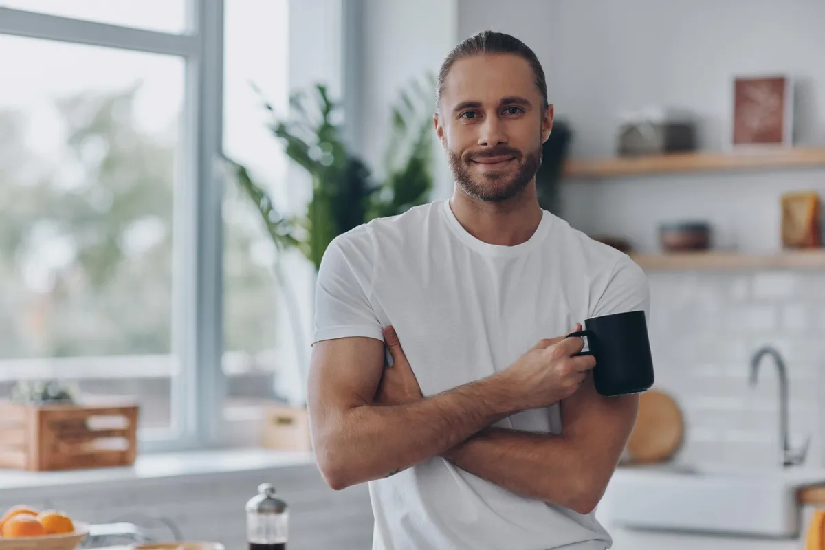 A man smiling in his kitchen while holding a coffee mug